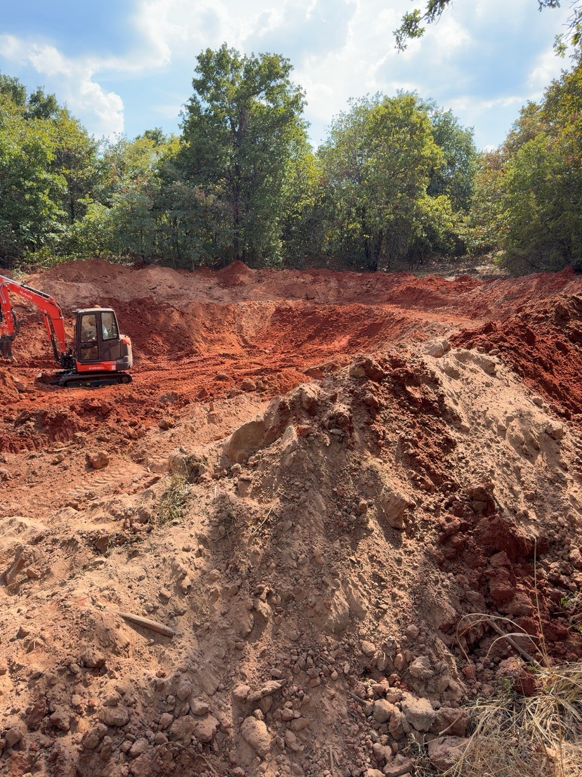 Pond excavation and red-clay earthmoving in progress beside woodland in Central Oklahoma